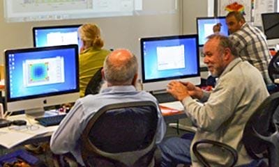 People are seated in a classroom with iMac computers, engaging in discussion and looking at projected content on a screen at the front of the room.
