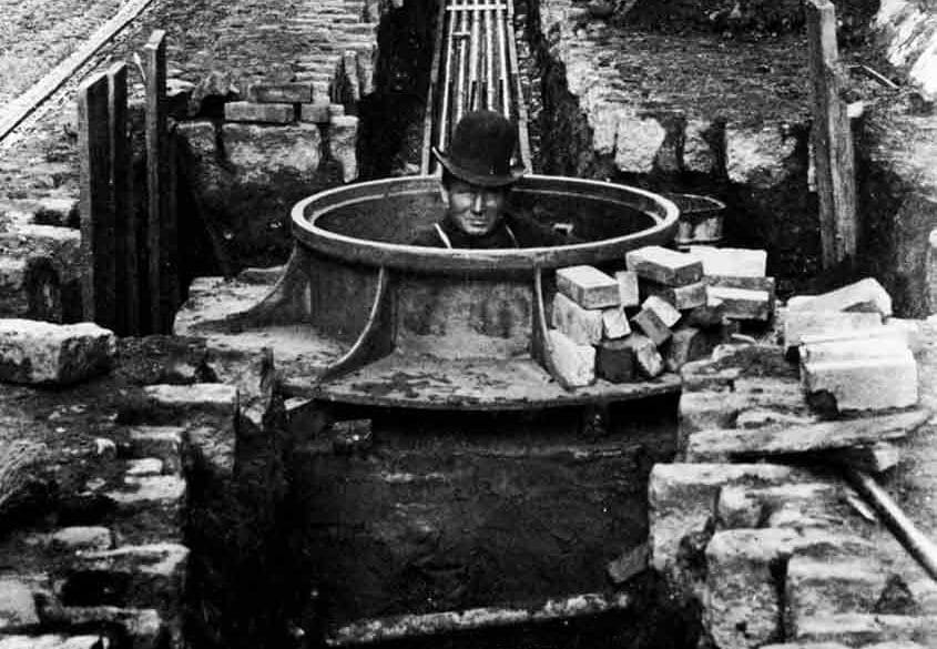 A worker in a manhole during early electrica power cable installation in New York city. An open trench and scattered bricks surround the area.