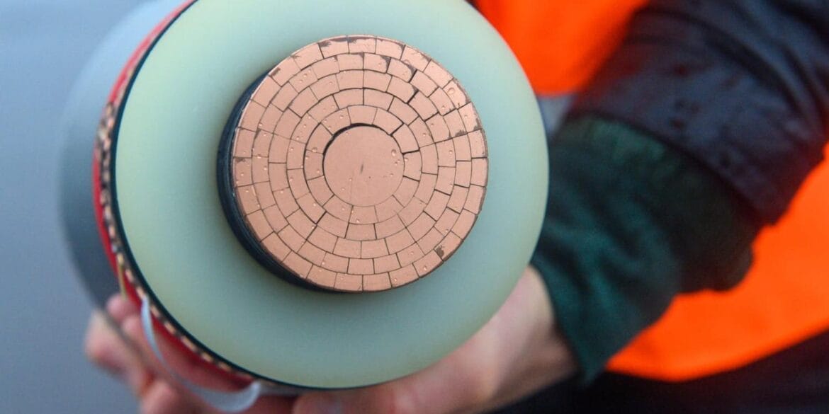 A person wearing an orange safety vest holds a cross-section of a high-voltage power cable, showing multiple concentric copper segments.