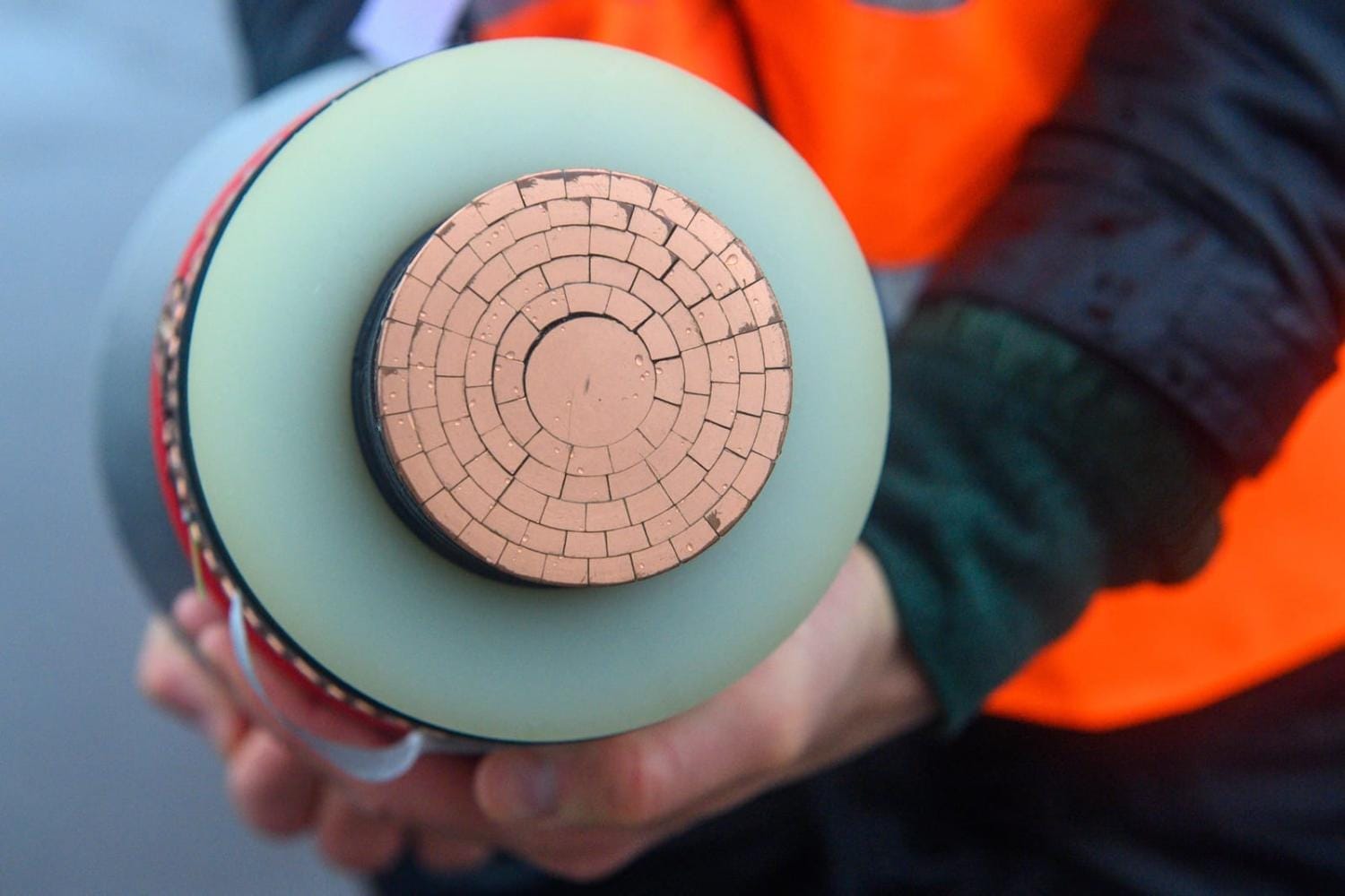 A person wearing an orange safety vest holds a cross-section of a high-voltage power cable, showing multiple concentric copper segments. A person wearing an orange safety vest holds a cross-section of a high-voltage power cable, showing multiple concentric copper segments.