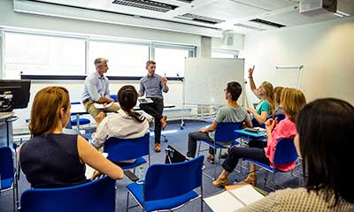 A group of people sit in a classroom facing two men at the front, one person raises their hand; a whiteboard is in the background.