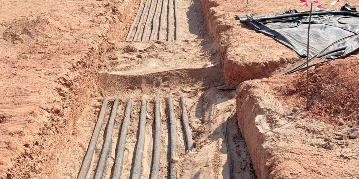 Several black cables are laid in a trench dug through dry soil at a construction site, with metal posts and flagged stakes visible in the background.
